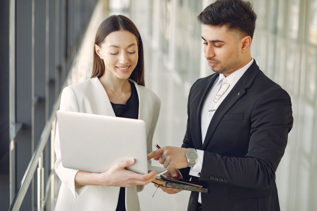 Concentrated diverse male and female employees in formal clothes standing in modern office corridor and browsing laptop while discussing business project together